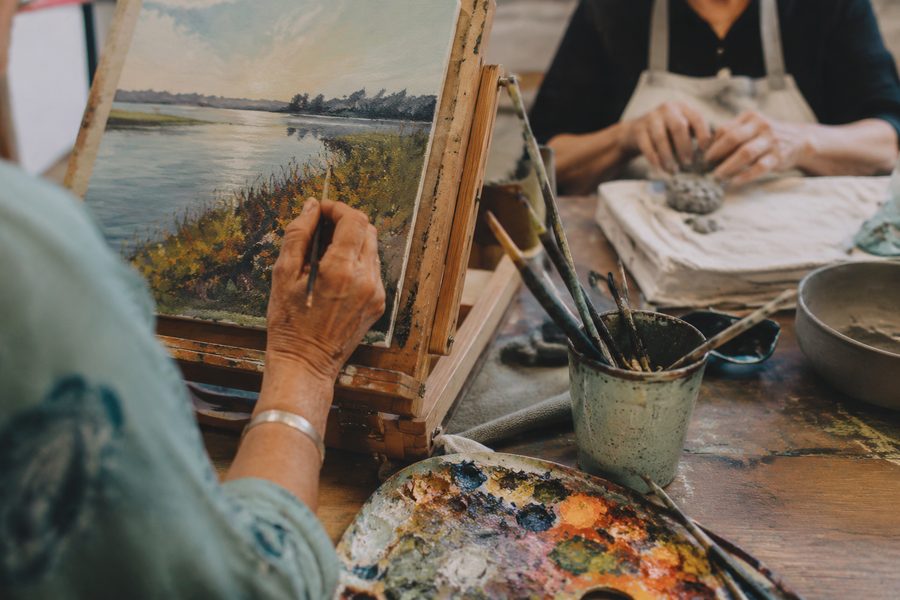 A participant paints a landscape at a table easel while another works with clay
